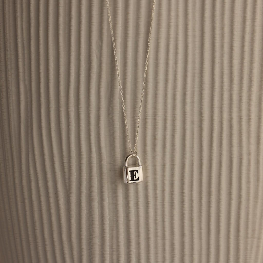 The Padlock Necklace features a silver chain with a small E-engraved pendant, offering a subtle and stylish fashion statement against a textured, light background.