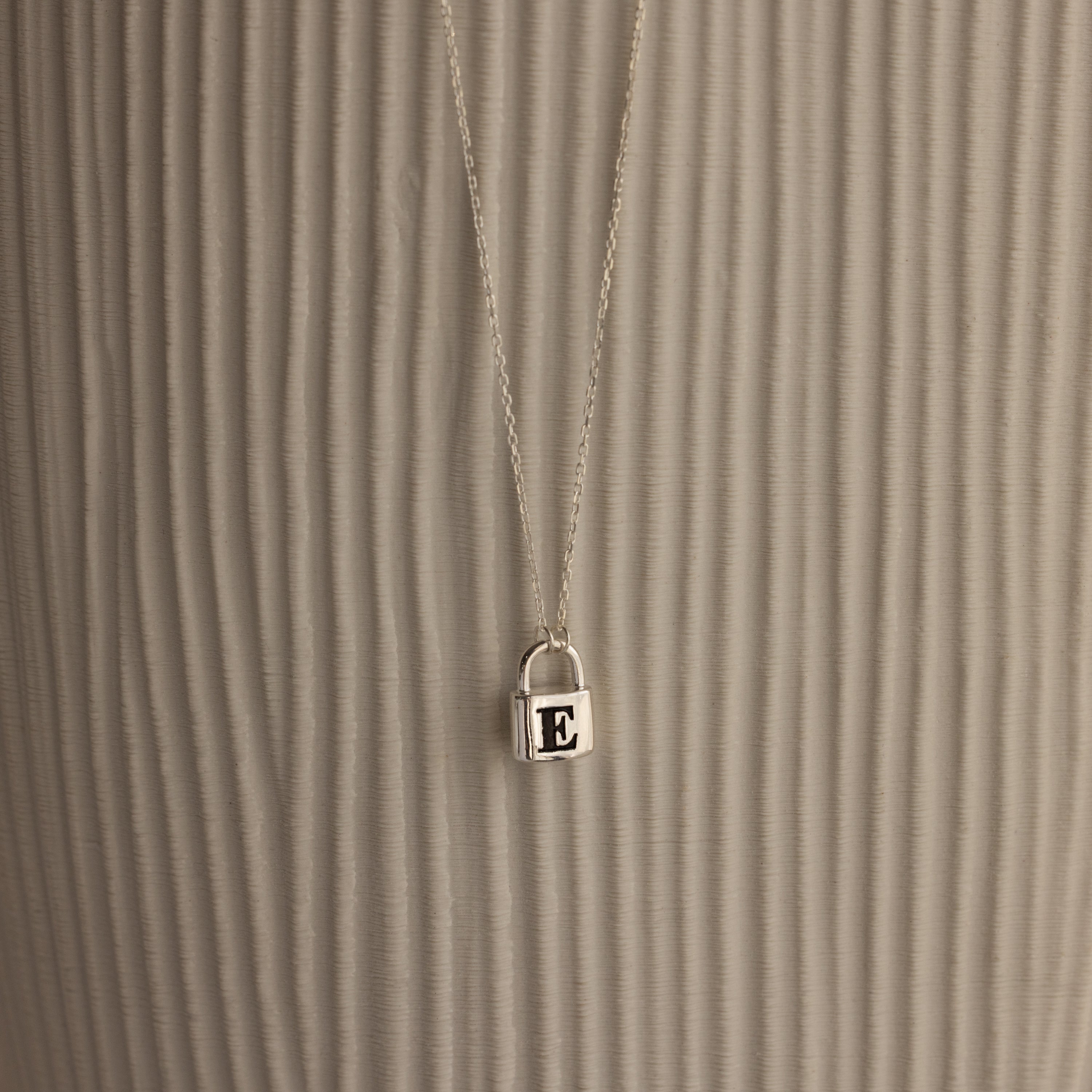 The Padlock Necklace features a silver chain with a small E-engraved pendant, offering a subtle and stylish fashion statement against a textured, light background.