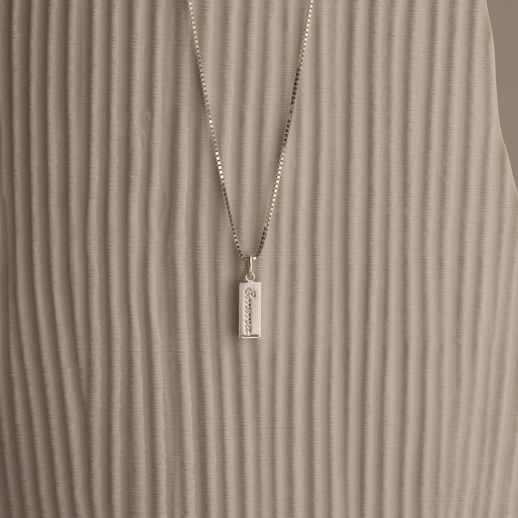 The Dainty Tag Name Necklace in Sterling Silver, featuring engraved text on a delicate chain, is displayed against a textured, light-colored background.