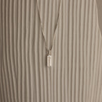The Dainty Tag Name Necklace in Sterling Silver, featuring engraved text on a delicate chain, is displayed against a textured, light-colored background.