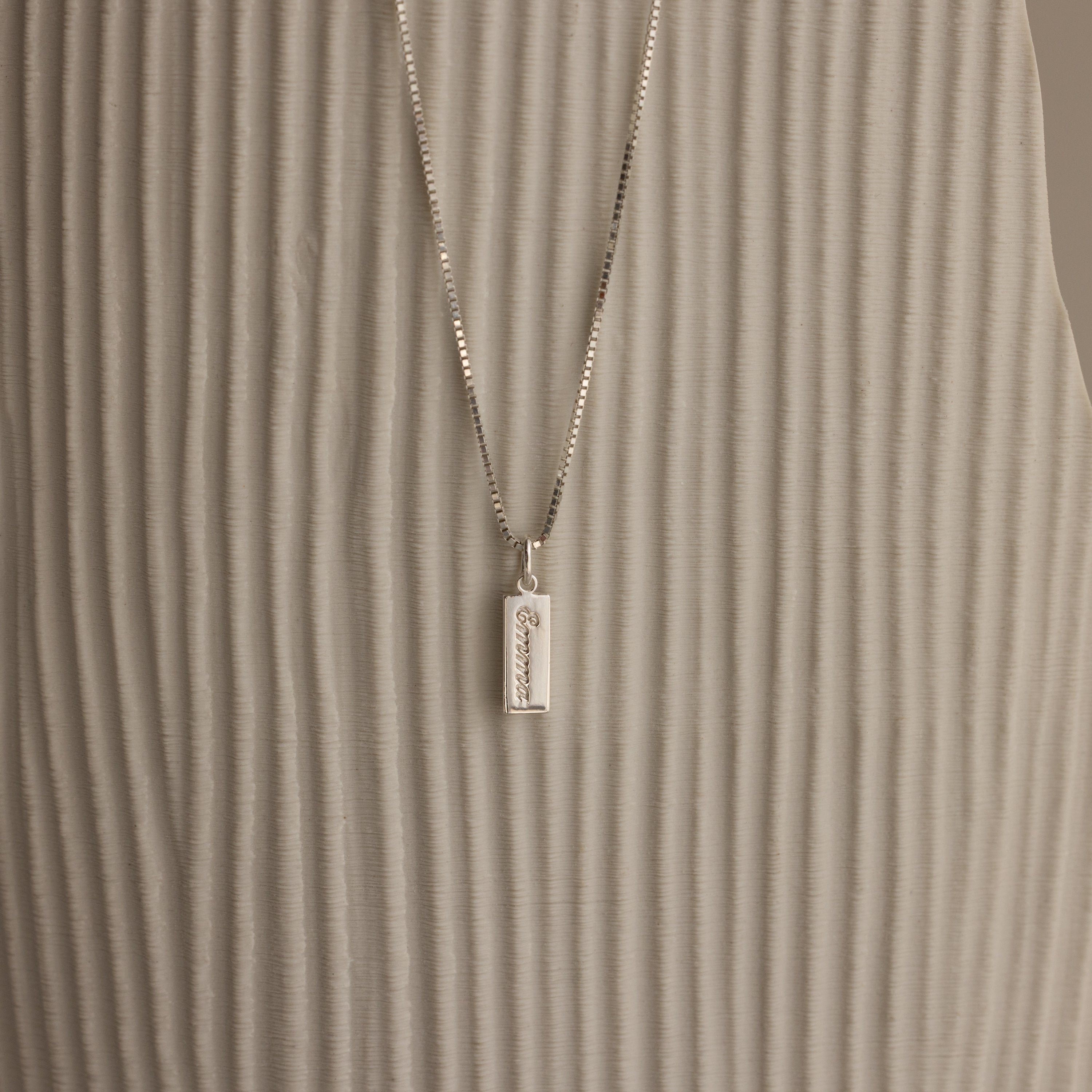 The Dainty Tag Name Necklace in Sterling Silver, featuring engraved text on a delicate chain, is displayed against a textured, light-colored background.