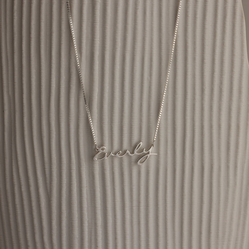 The Callie Box Chain Name Necklace features "Everly" in cursive on a delicate chain, shown against a textured, light background.