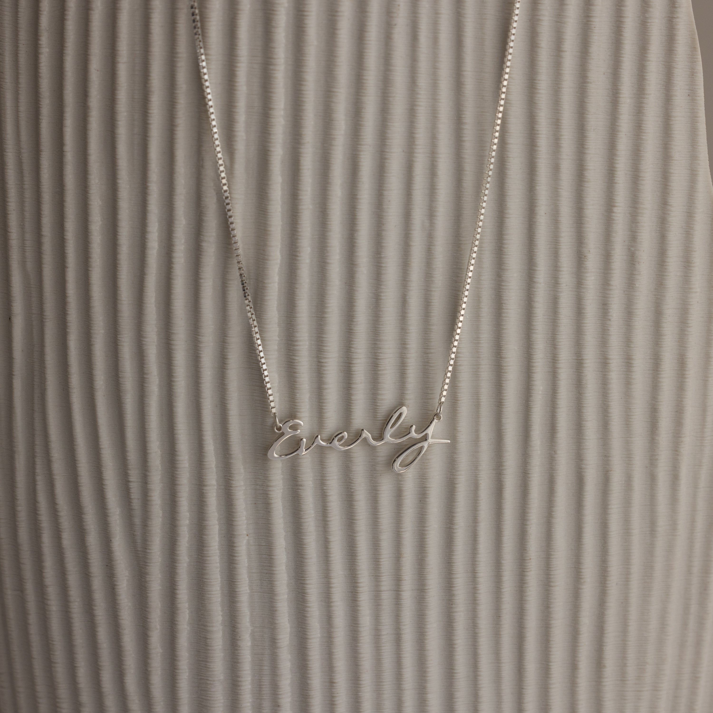 The Callie Box Chain Name Necklace features "Everly" in cursive on a delicate chain, shown against a textured, light background.