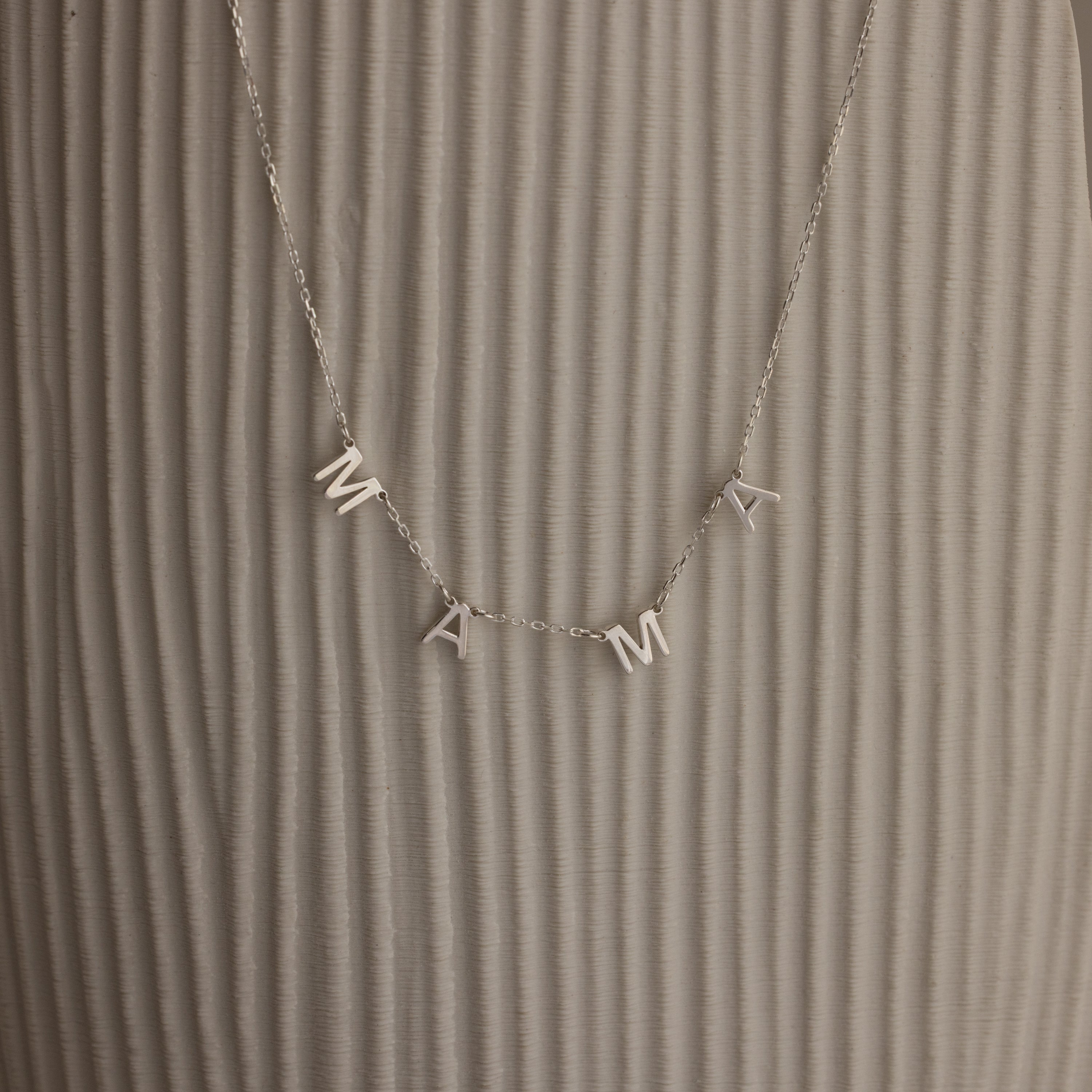 The Alice Letter Name Necklace in Sterling Silver with MAMA charms is displayed on a textured beige background—a perfect personalized necklace and thoughtful Christmas gift.