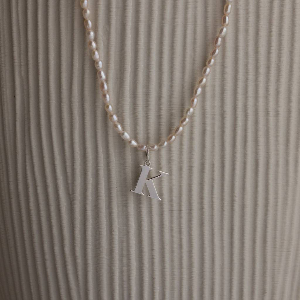 The Newsletter Pearl Initial Necklace in Sterling Silver features a sleek letter K pendant with pearl accents, displayed against a textured beige background—ideal for fans of minimalist jewelry.