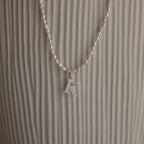 The Newsletter Pearl Initial Necklace in Sterling Silver features a sleek letter K pendant with pearl accents, displayed against a textured beige background—ideal for fans of minimalist jewelry.