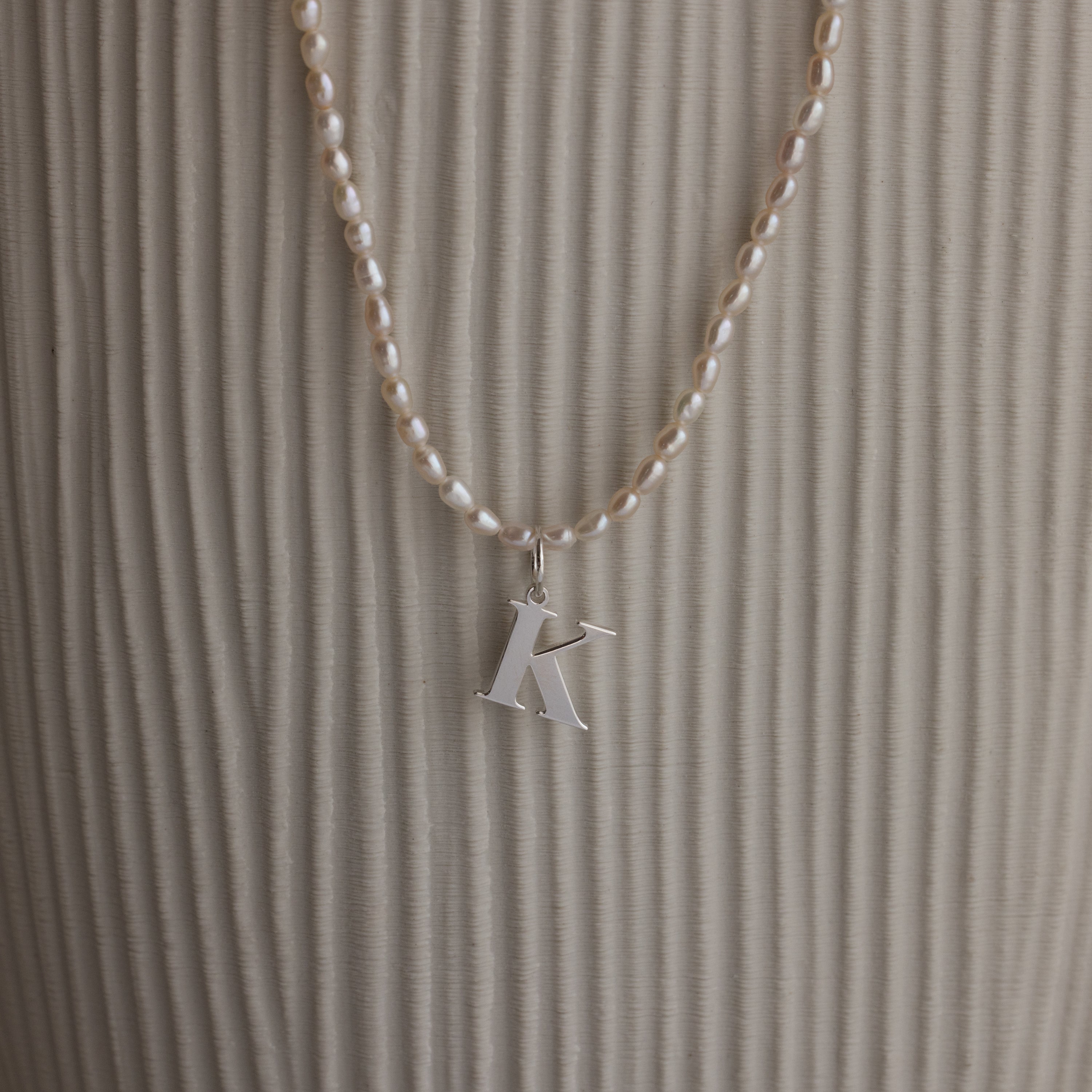 The Newsletter Pearl Initial Necklace in Sterling Silver features a sleek letter K pendant with pearl accents, displayed against a textured beige background—ideal for fans of minimalist jewelry.