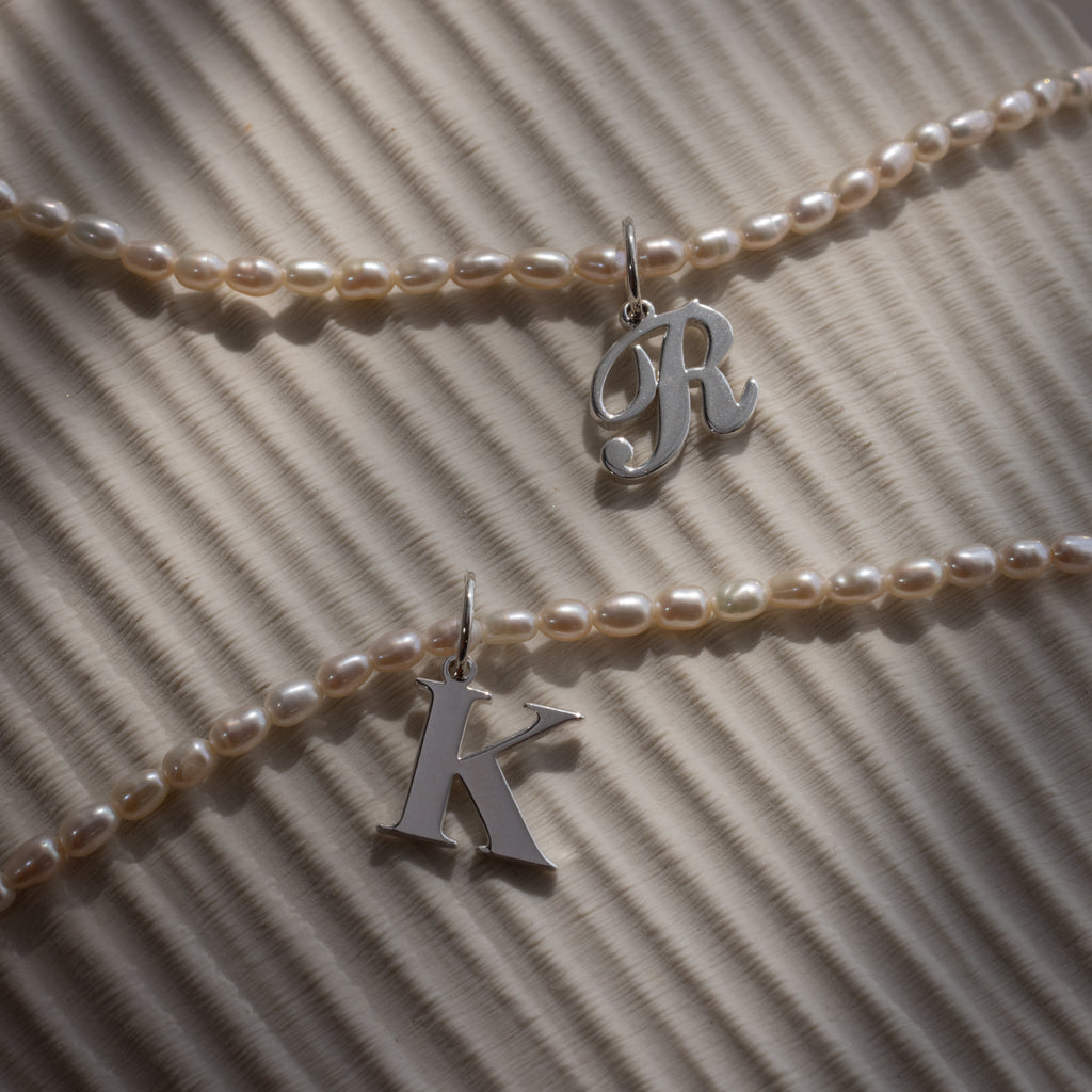 Two Newsletter Pearl Initial Necklaces in Sterling Silver, featuring customizable letter charms R and K, rest on a striped, sunlit background—ideal for fans of minimalist jewelry with personal flair.