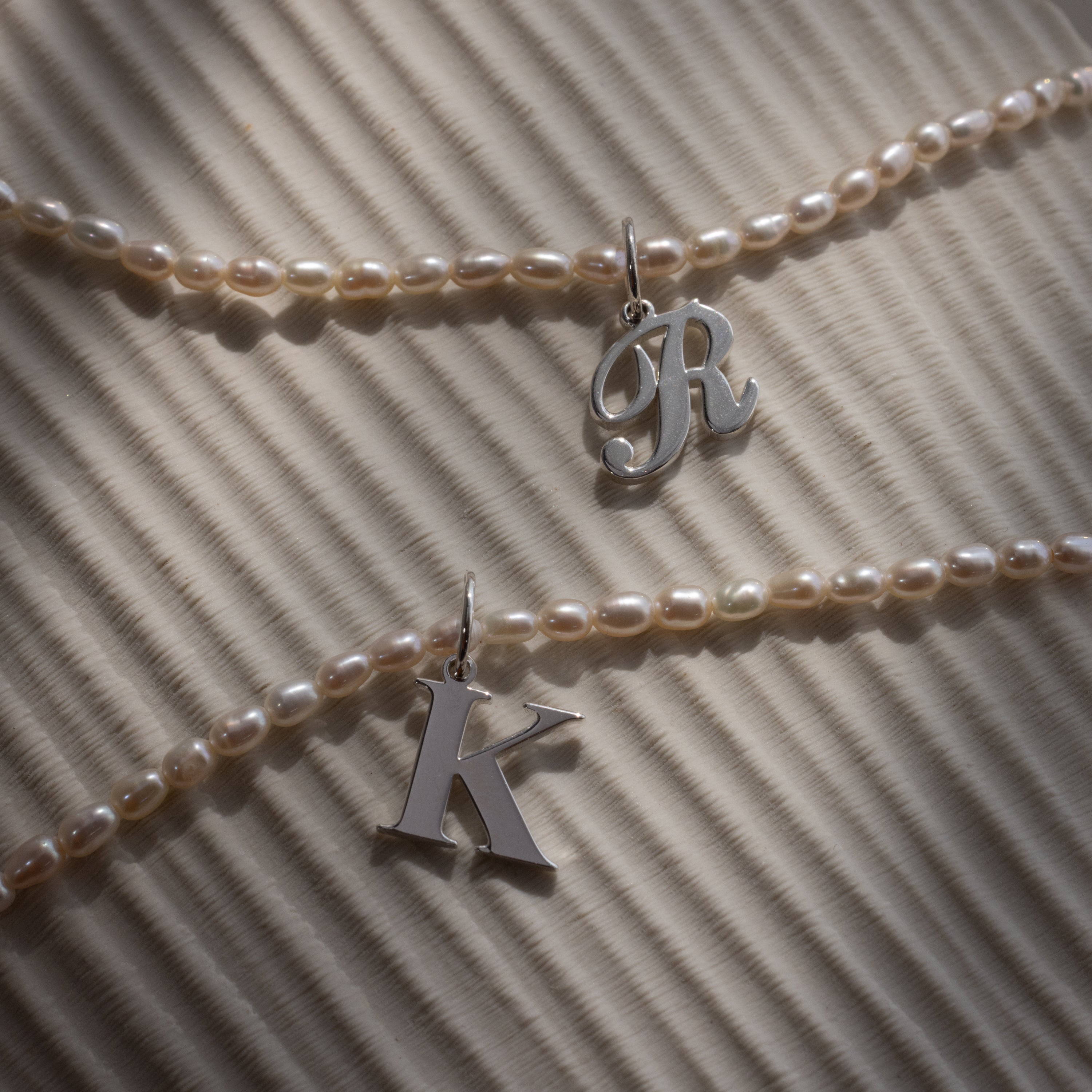 Two Newsletter Pearl Initial Necklaces in Sterling Silver, featuring customizable letter charms R and K, rest on a striped, sunlit background—ideal for fans of minimalist jewelry with personal flair.