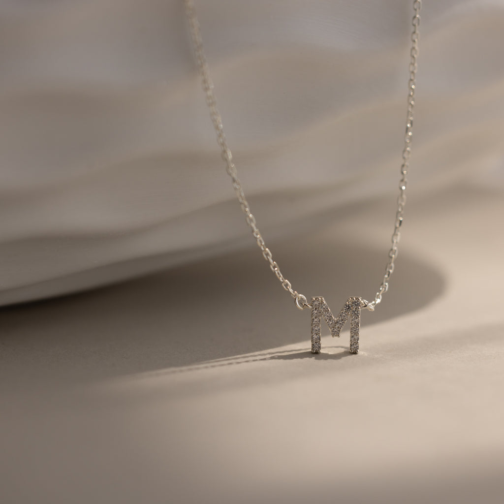 The Pave Initial Necklace features a sparkling, diamond-studded letter M pendant in silver, displayed on a light textured surface—a refined and personalized jewelry piece.