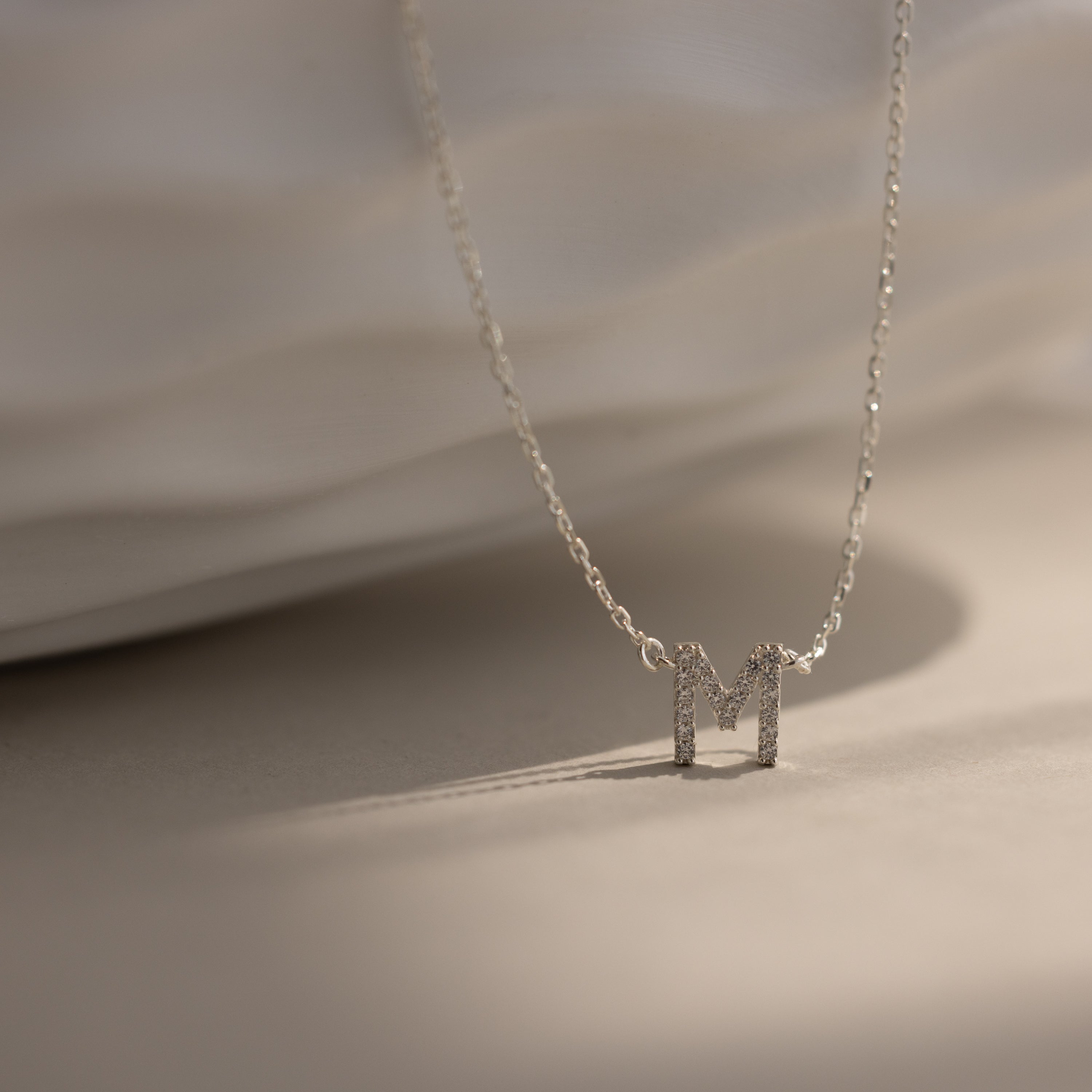 The Pave Initial Necklace features a sparkling, diamond-studded letter M pendant in silver, displayed on a light textured surface—a refined and personalized jewelry piece.