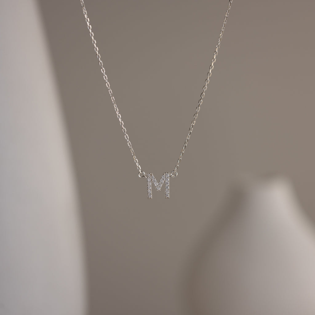 The Pave Initial Necklace features a sparkling letter M pendant, adding personalized elegance to your look against a blurred neutral background.
