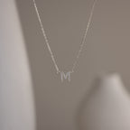 The Pave Initial Necklace features a sparkling letter M pendant, adding personalized elegance to your look against a blurred neutral background.
