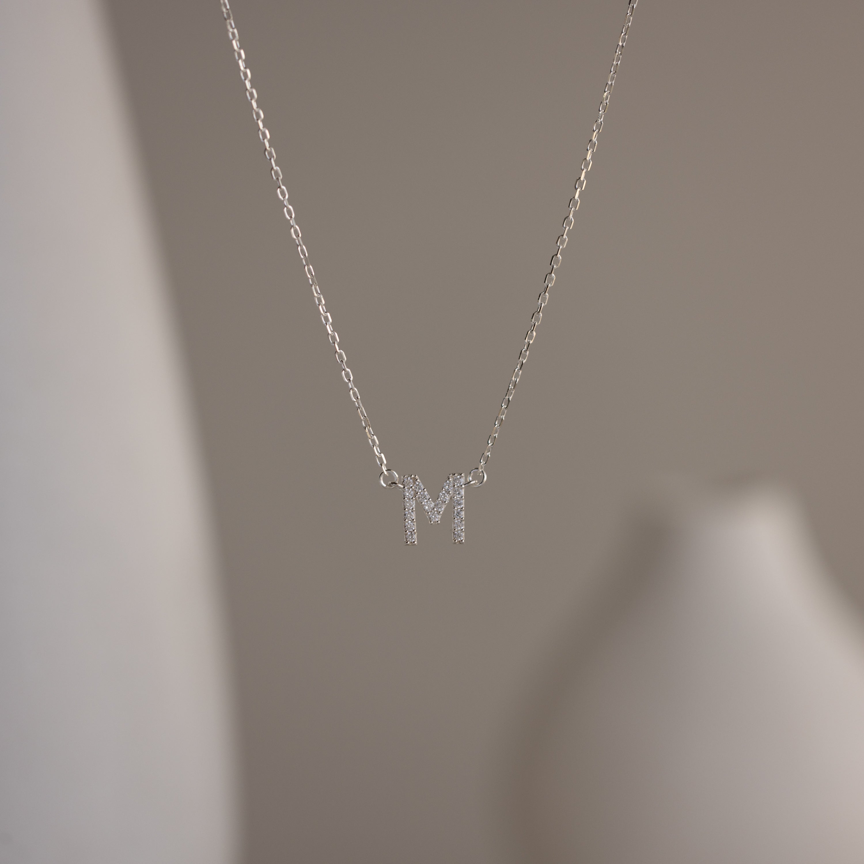 The Pave Initial Necklace features a sparkling letter M pendant, adding personalized elegance to your look against a blurred neutral background.
