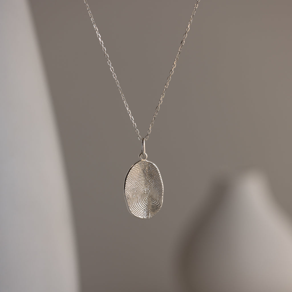 The Custom Fingerprint Necklace features a silver oval fingerprint pendant, displayed against a blurred neutral background to highlight this elegant piece of personalized jewelry.
