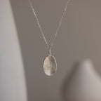 The Custom Fingerprint Necklace features a silver oval fingerprint pendant, displayed against a blurred neutral background to highlight this elegant piece of personalized jewelry.