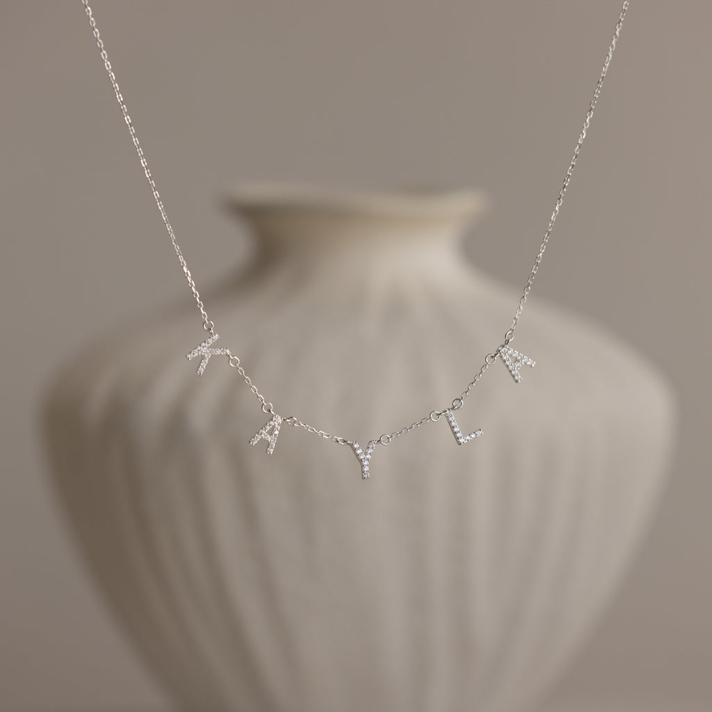 The Pave Letter Necklace in Sterling Silver, featuring charms that spell "KYL," is displayed against a blurred white vase, showcasing personalized elegance.