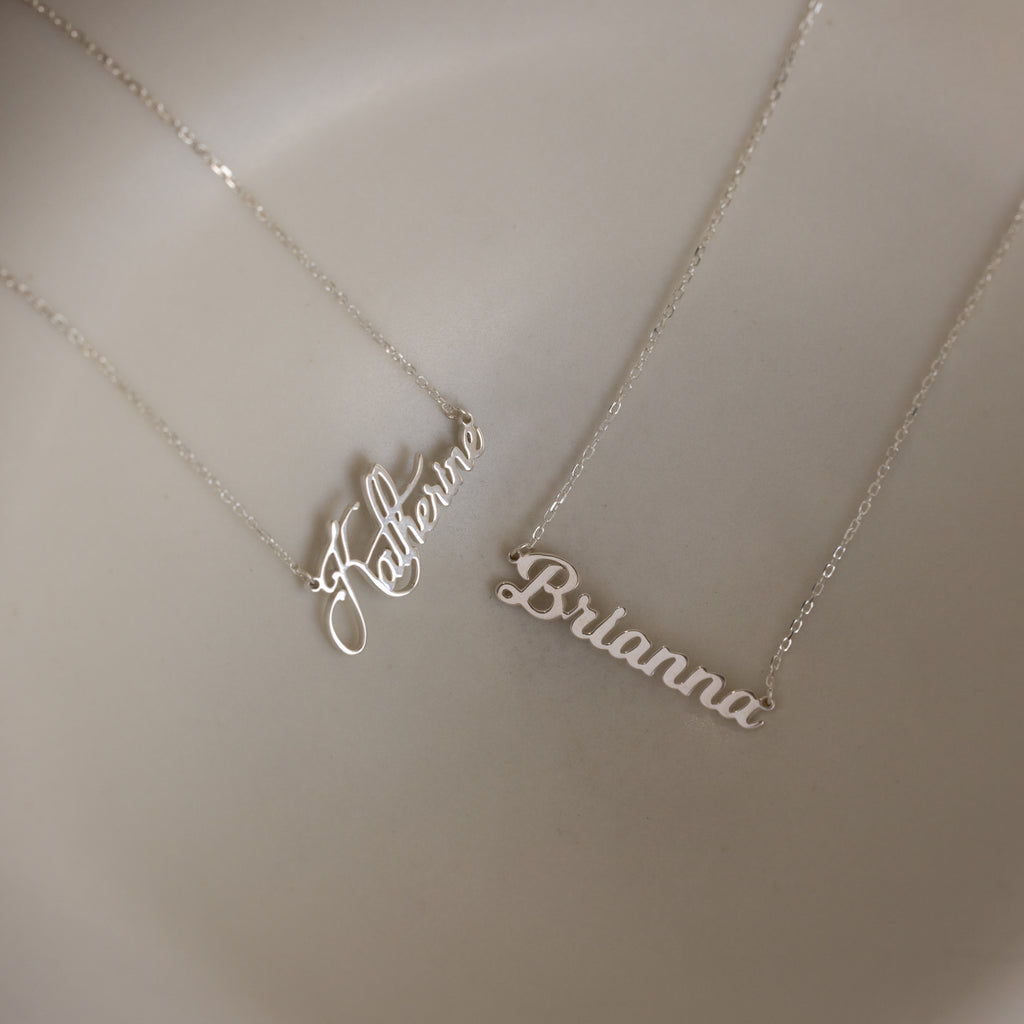 Two Mellow Name Necklaces in Sterling Silver rest on a beige surface, each featuring a unique script—one spelling Katherine and the other Brianna. Personalize your own Mellow Name Necklace for a custom look.