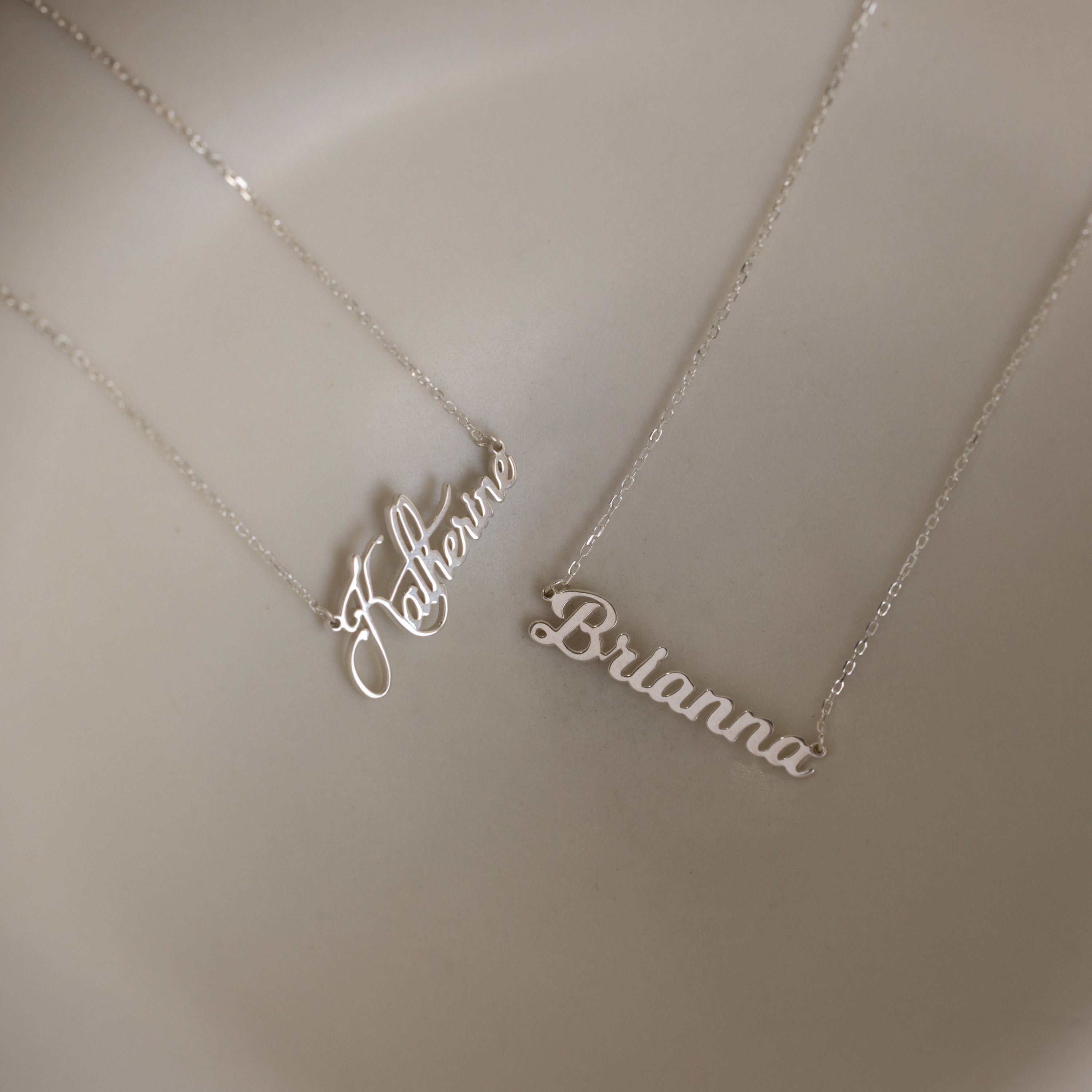 Two Mellow Name Necklaces in Sterling Silver rest on a beige surface, each featuring a unique script—one spelling Katherine and the other Brianna. Personalize your own Mellow Name Necklace for a custom look.