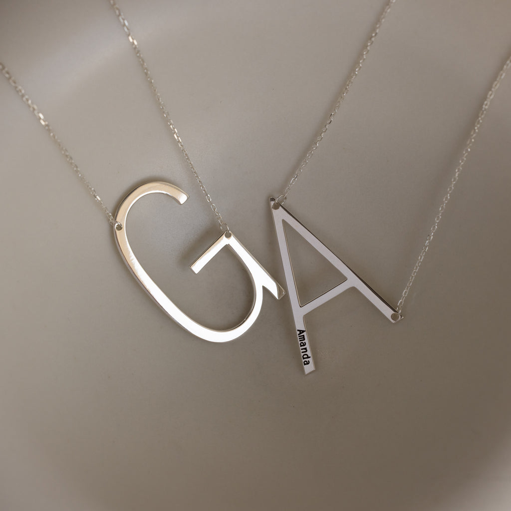 Two Large Sideways Initial Necklaces in Sterling Silver with bold letter pendants "G" and "A" rest on a smooth beige background, highlighting the striking design of this personalized jewelry.