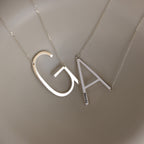 Two Large Sideways Initial Necklaces in Sterling Silver with bold letter pendants "G" and "A" rest on a smooth beige background, highlighting the striking design of this personalized jewelry.