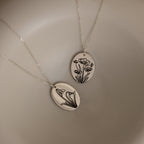 The Oval Flower Necklace in Sterling Silver features two silver oval pendants with delicate floral engravings on fine chains—perfect as personalized or birth flower jewelry. Shown on a neutral background.