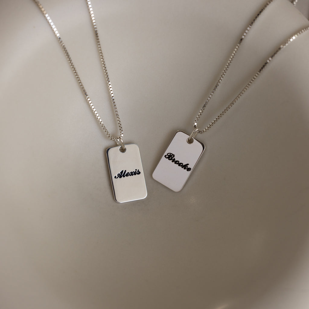 Two Name Tag Necklaces in Sterling Silver with rectangular pendants showcase personalized engraving—elegant jewelry featuring the names "Alexis" and "Brooke" on a sleek, plain surface.