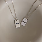 Two Name Tag Necklaces in Sterling Silver with rectangular pendants showcase personalized engraving—elegant jewelry featuring the names "Alexis" and "Brooke" on a sleek, plain surface.