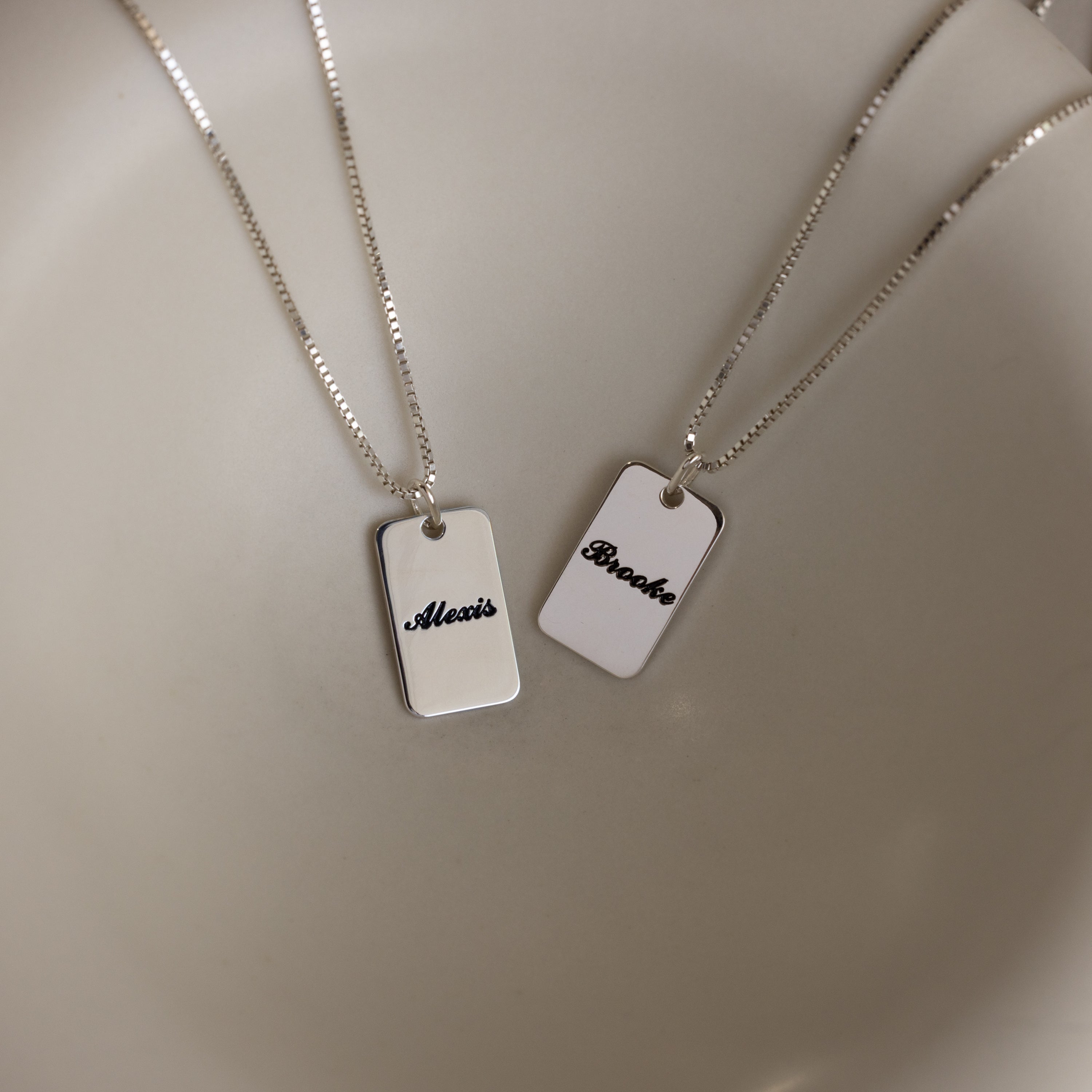 Two Name Tag Necklaces in Sterling Silver with rectangular pendants showcase personalized engraving—elegant jewelry featuring the names "Alexis" and "Brooke" on a sleek, plain surface.