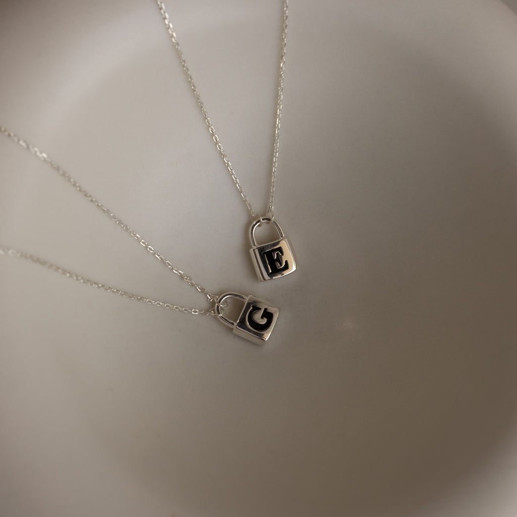 Two Padlock Necklaces in Sterling Silver, each with an engraved initial—one E, one G—rest on a white surface, creating a bold fashion statement.