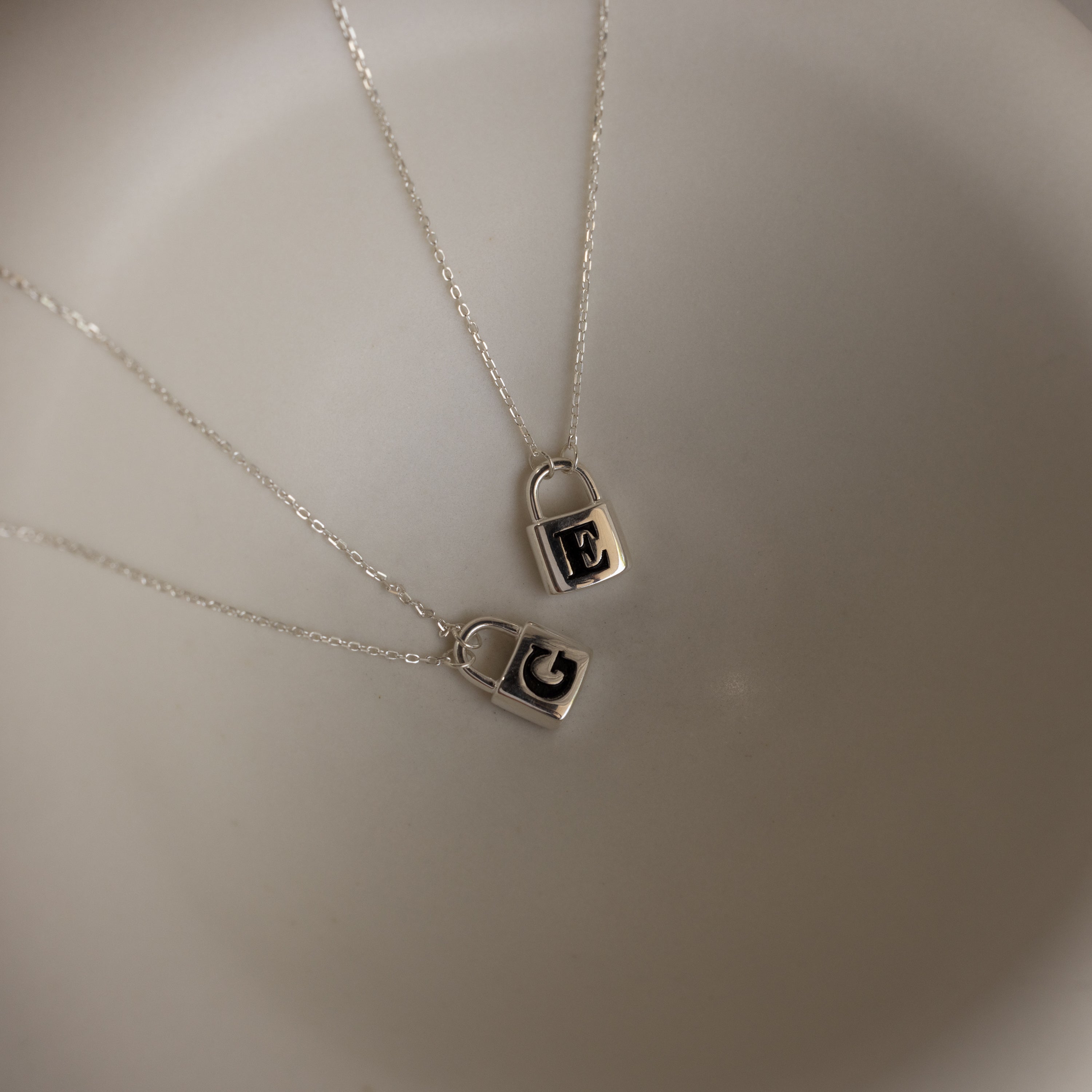 Two Padlock Necklaces in Sterling Silver, each with an engraved initial—one E, one G—rest on a white surface, creating a bold fashion statement.