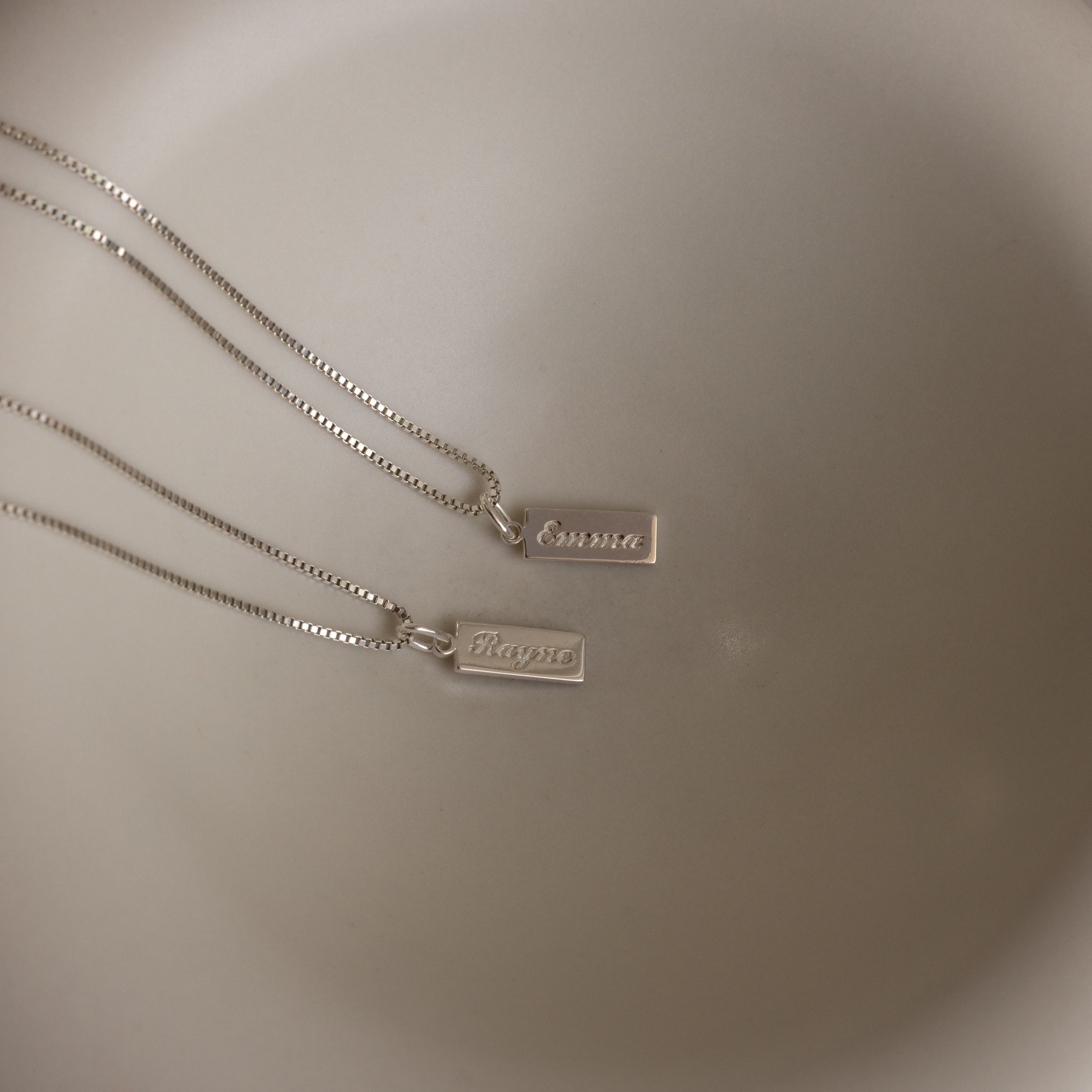 Two Dainty Tag Name Necklaces in Sterling Silver with rectangular pendants engraved "Emma" and "Magnu" hang from delicate chains against a neutral background.