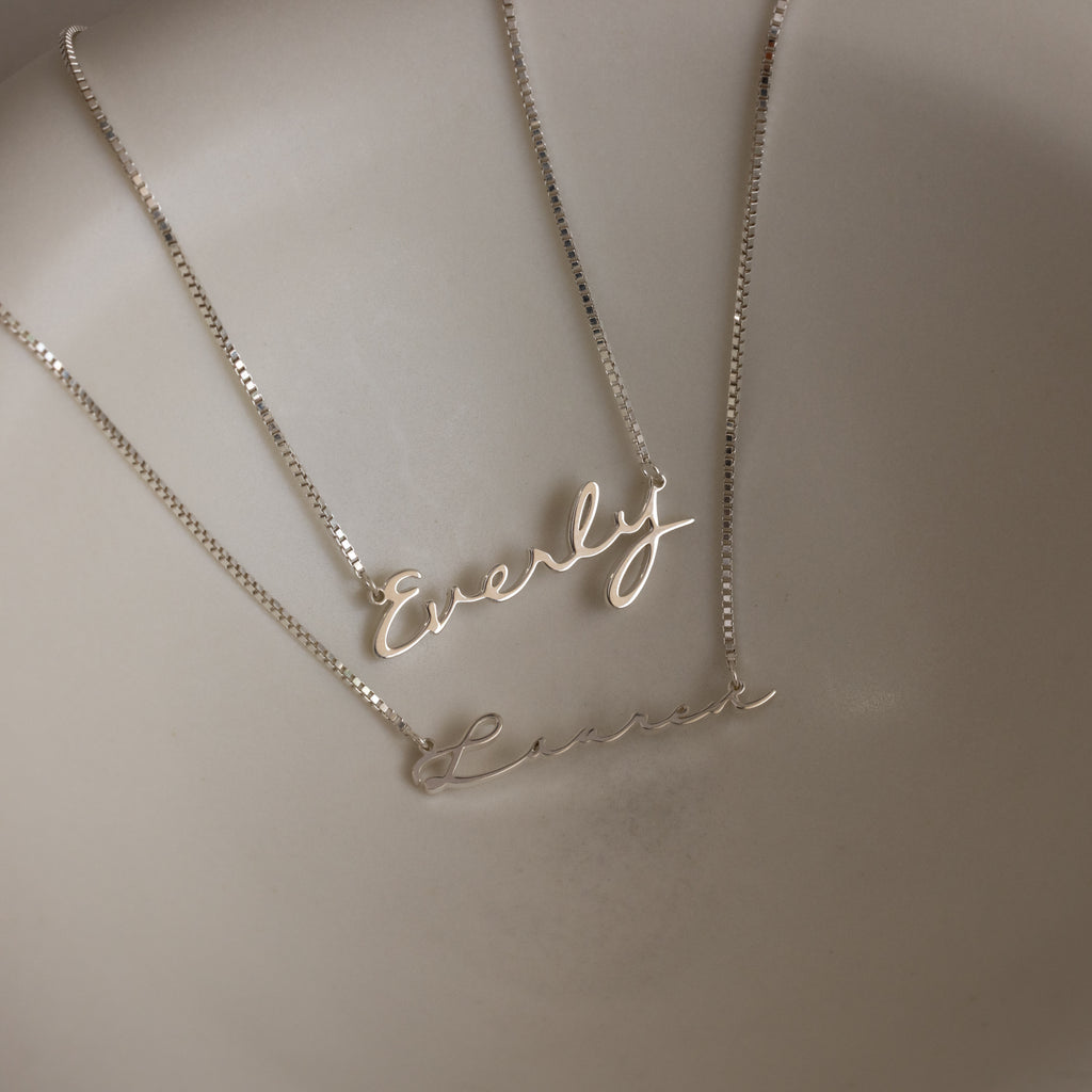 Two Callie Box Chain Name Necklaces in Sterling Silver, each with elegant cursive script on a delicate chain, are displayed on a smooth, cream-colored surface—ideal as customizable charms to suit any style.