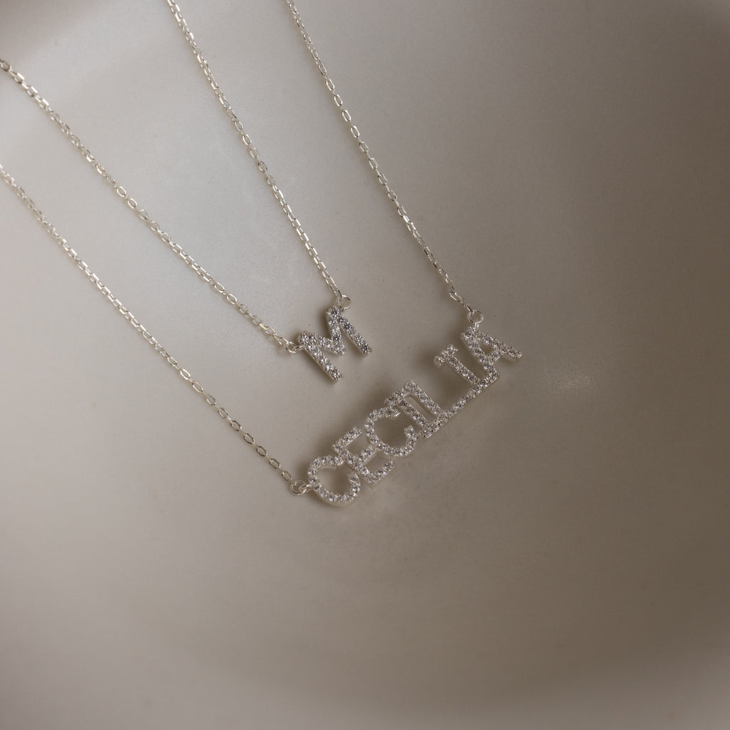 Two delicate sterling silver necklaces, including a Pave Initial Necklace with an "M" pendant and a personalized necklace spelling out "CECILIA," are displayed on a neutral background.