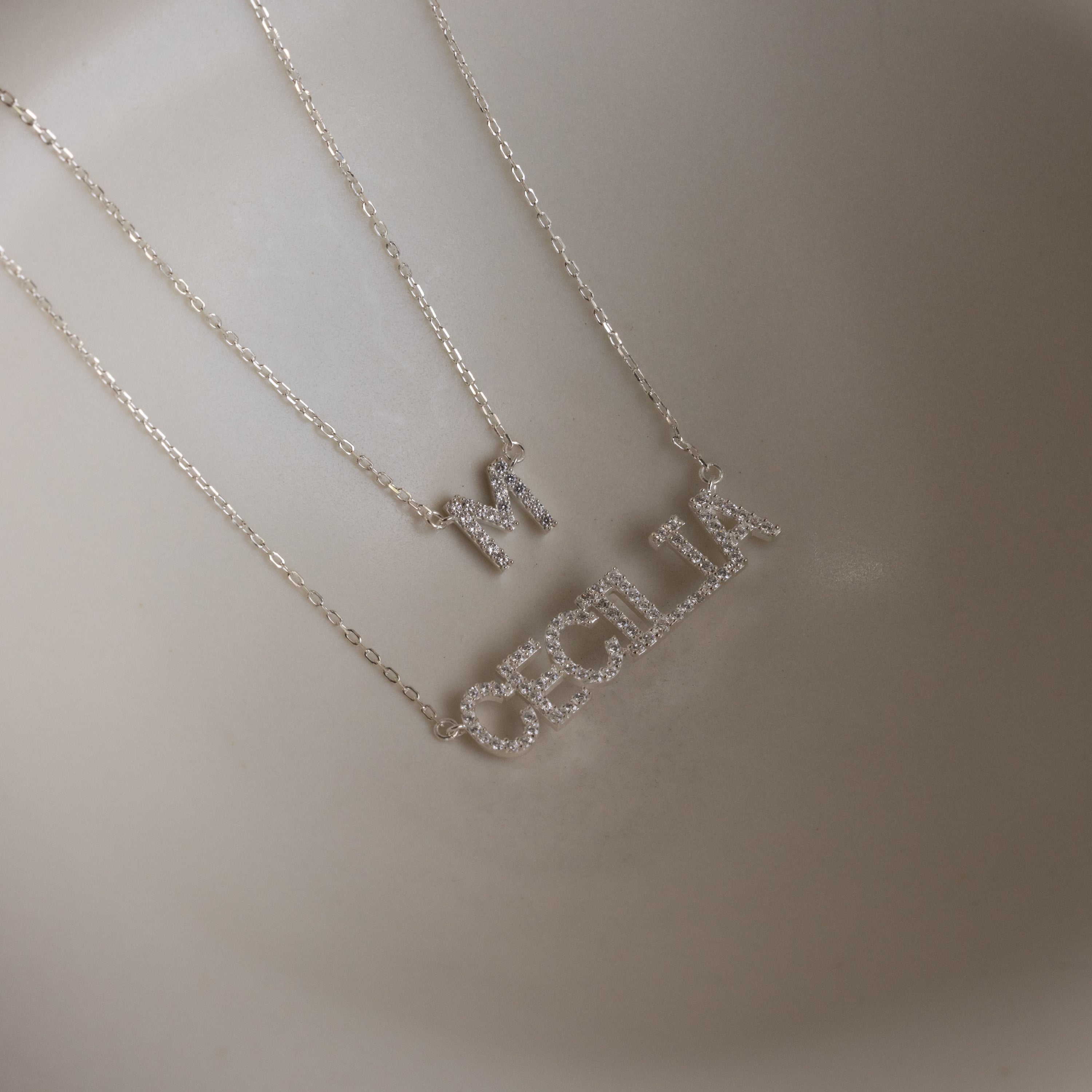 Two delicate sterling silver necklaces, including a Pave Initial Necklace with an "M" pendant and a personalized necklace spelling out "CECILIA," are displayed on a neutral background.