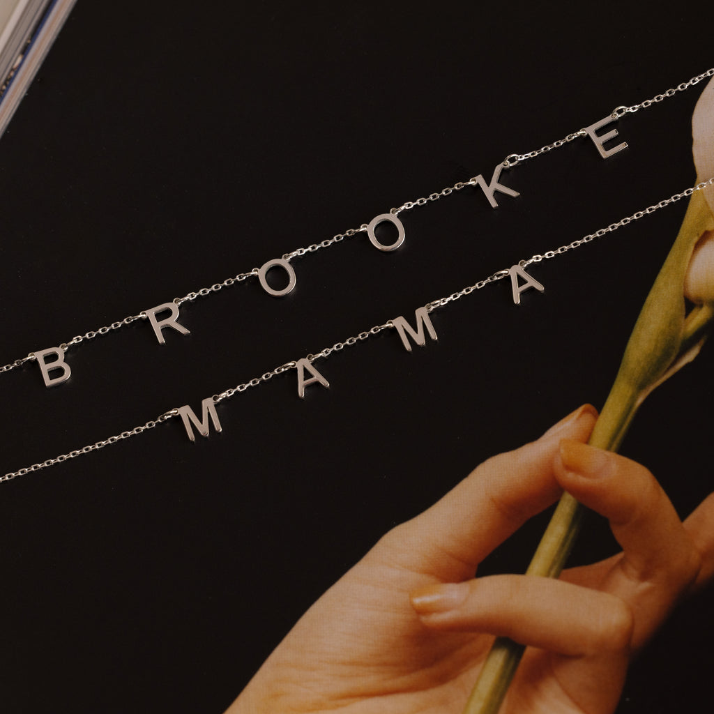 The Alice Letter Name Necklace in Sterling Silver spells out names like BROOKE and MAMA, shown beside a pale flower on a dark background—a stylish personalized necklace, ideal for Christmas gifting.