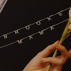 The Alice Letter Name Necklace in Sterling Silver spells out names like BROOKE and MAMA, shown beside a pale flower on a dark background—a stylish personalized necklace, ideal for Christmas gifting.