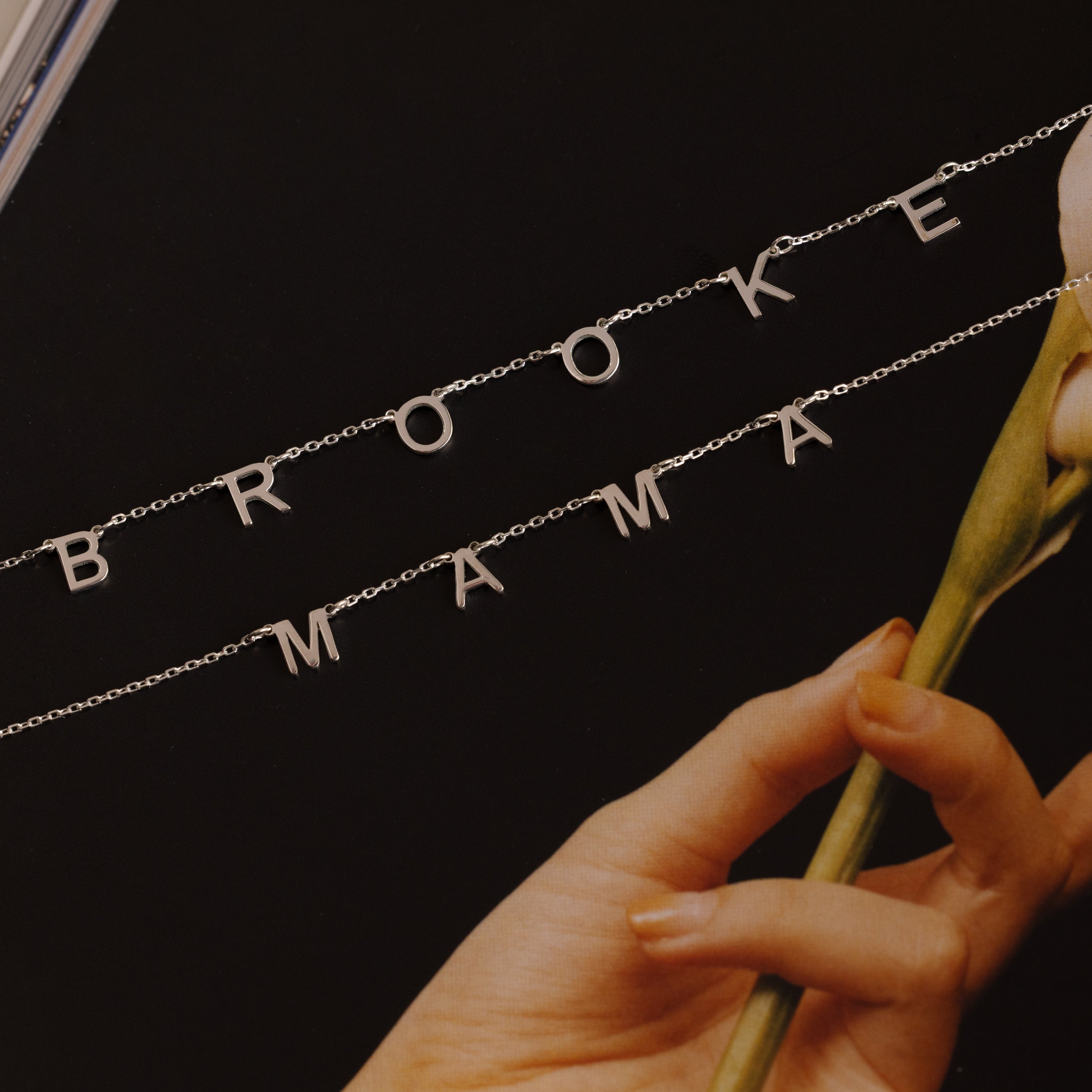 The Alice Letter Name Necklace in Sterling Silver spells out names like BROOKE and MAMA, shown beside a pale flower on a dark background—a stylish personalized necklace, ideal for Christmas gifting.