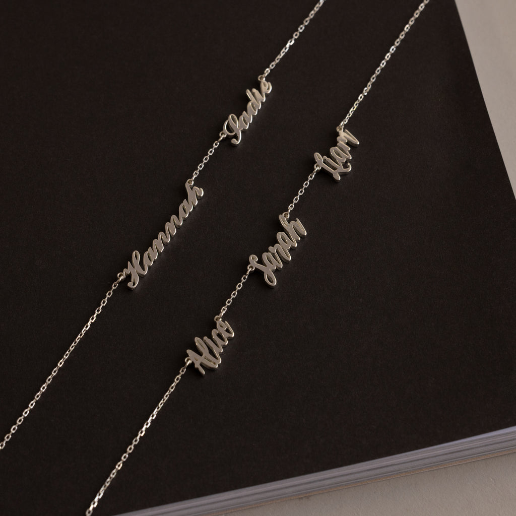Two Carrie Multiple Name Necklaces with silver cursive pendants reading “Sarah Leah” and “Mia Sarah Ilan” rest elegantly on a dark notebook, highlighting the charm of personalized jewelry.