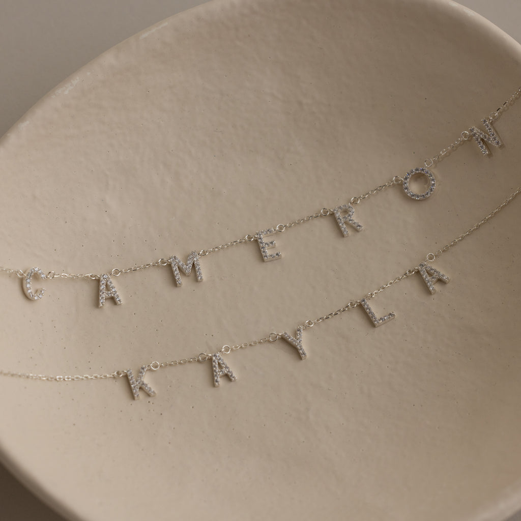 Two Pave Letter Necklaces in Sterling Silver rest on a cream dish, each with letter charms spelling CAMERON and KAYLA—an ideal choice for fans of delicate jewelry.