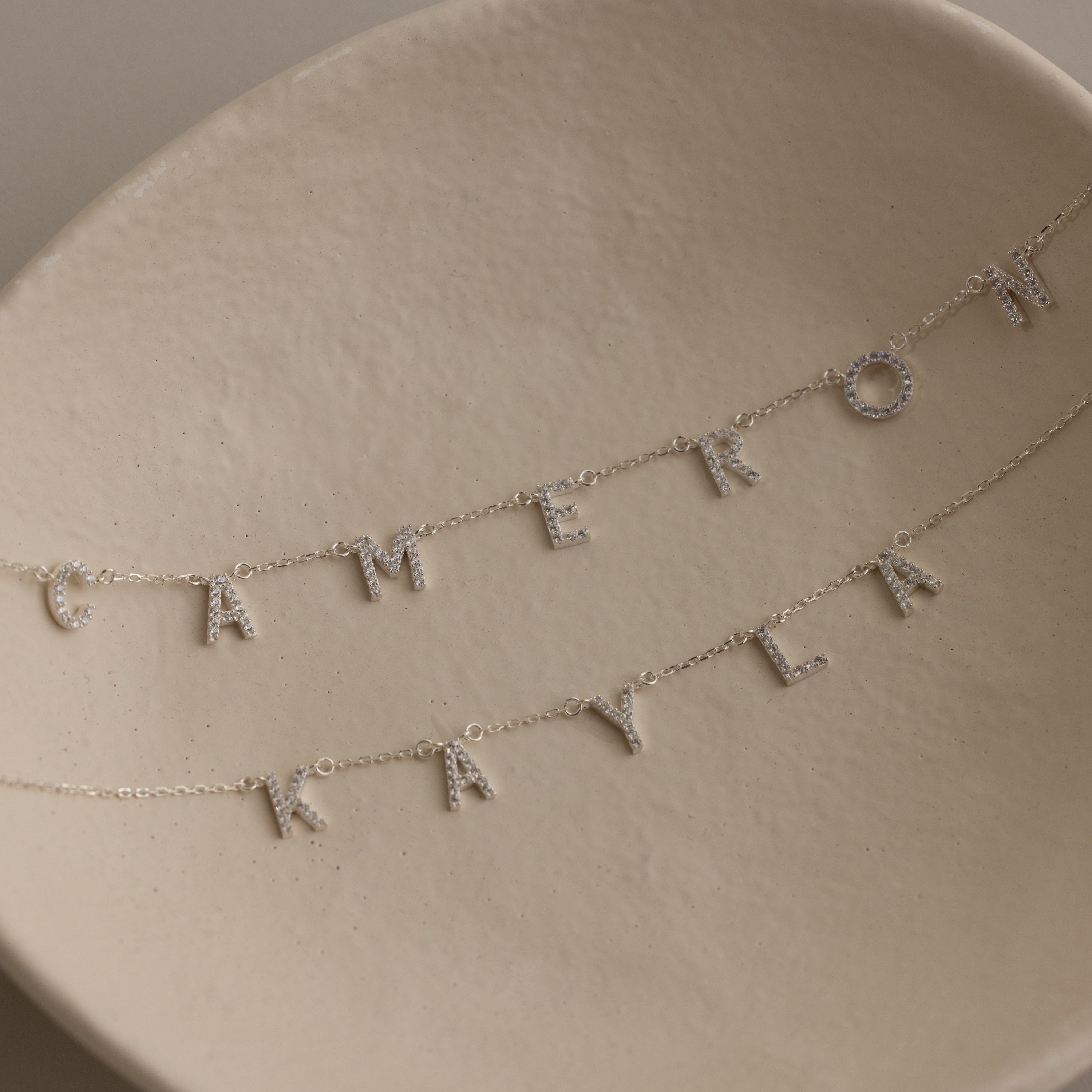 Two Pave Letter Necklaces in Sterling Silver rest on a cream dish, each with letter charms spelling CAMERON and KAYLA—an ideal choice for fans of delicate jewelry.