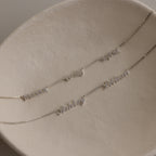 Two silver necklaces with multiple small name charm pendants on a beige surface