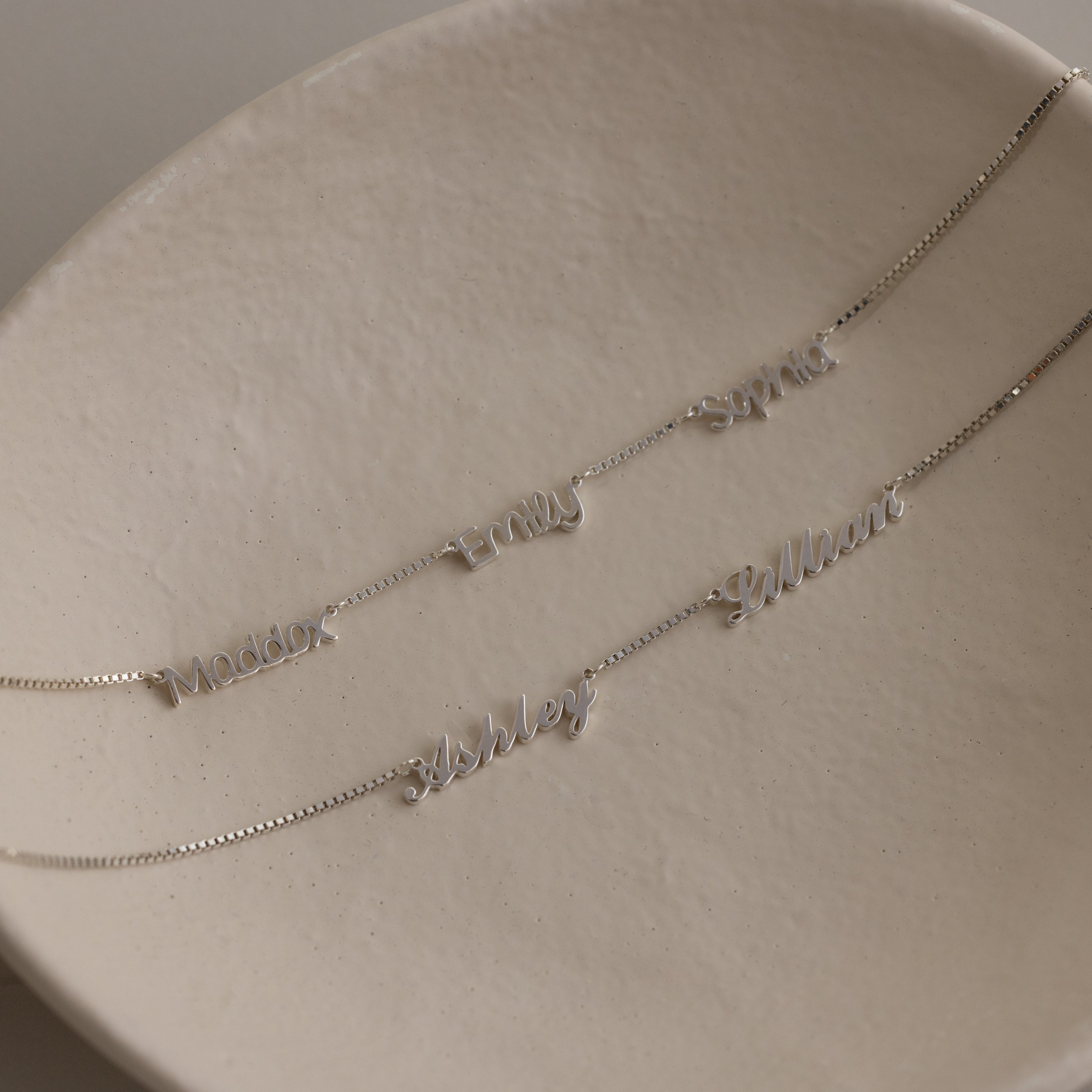 Two silver necklaces with multiple small name charm pendants on a beige surface