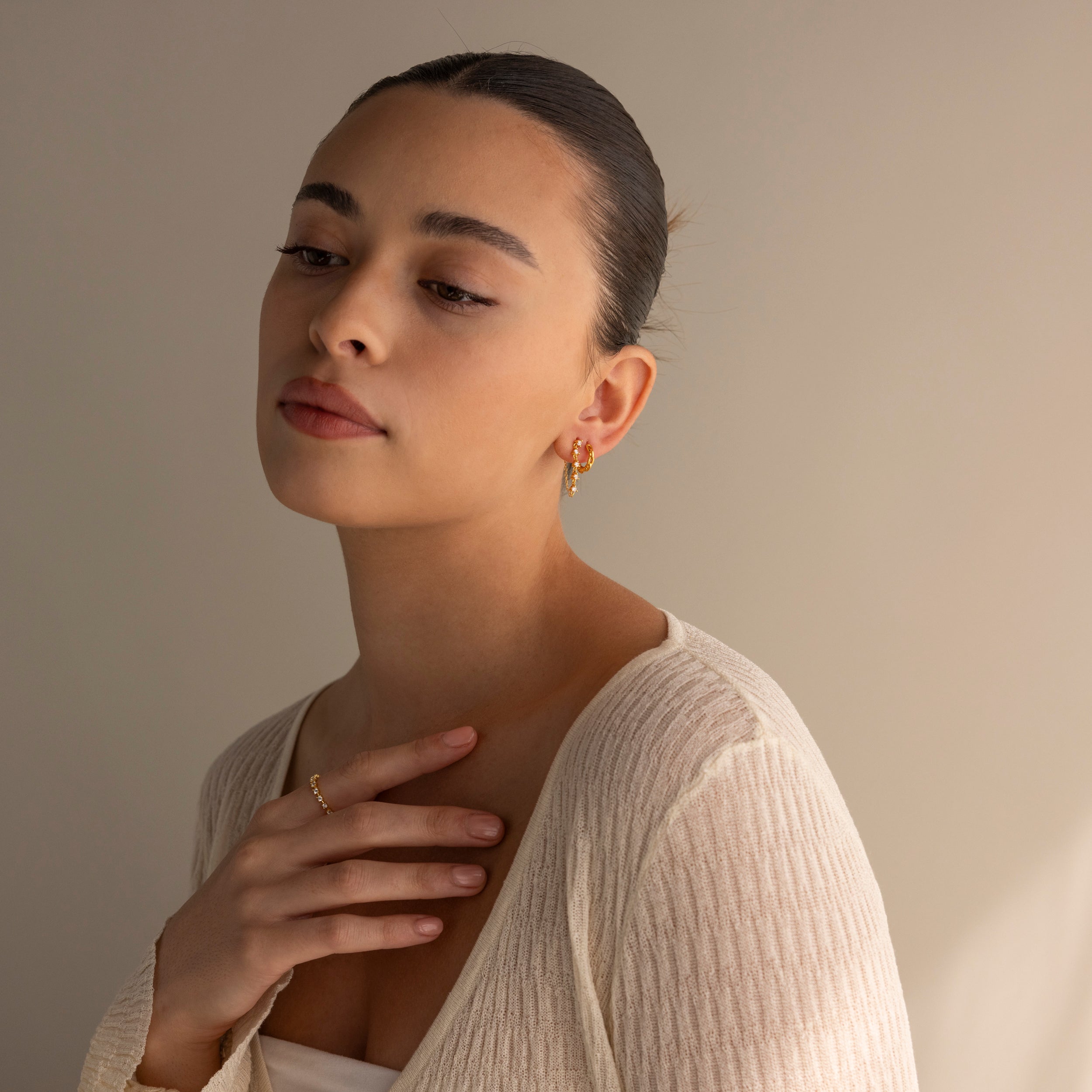 A woman with slicked-back hair and a cream textured top gazes downward thoughtfully, showcasing elegant Diamond Chain Drop Earrings.