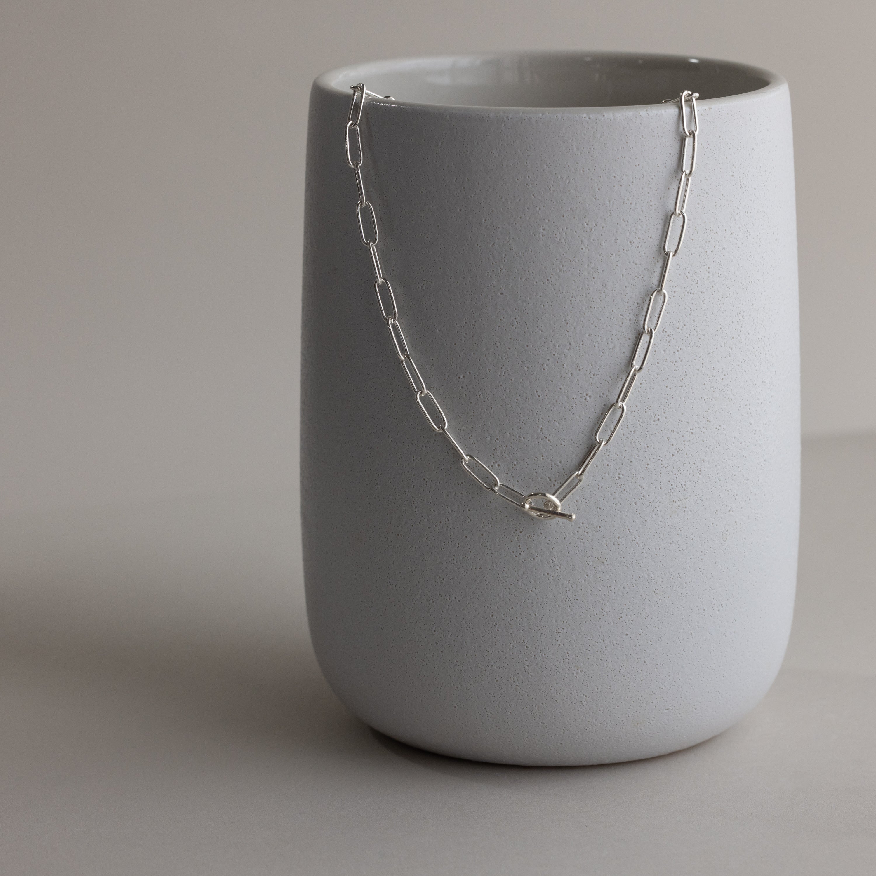 The Paperclip Toggle Necklace in Sterling Silver is displayed draped over a plain, matte gray ceramic cup.
