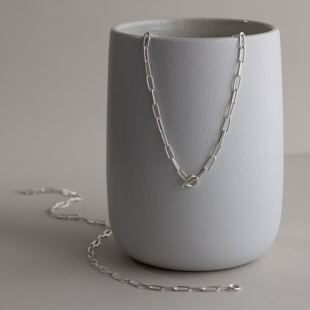 The Paperclip Toggle Necklace shines in silver atop a matte gray ceramic cup, bringing boho-chic elegance and versatility to your layered necklaces collection.
