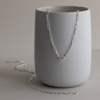 The Paperclip Toggle Necklace shines in silver atop a matte gray ceramic cup, bringing boho-chic elegance and versatility to your layered necklaces collection.