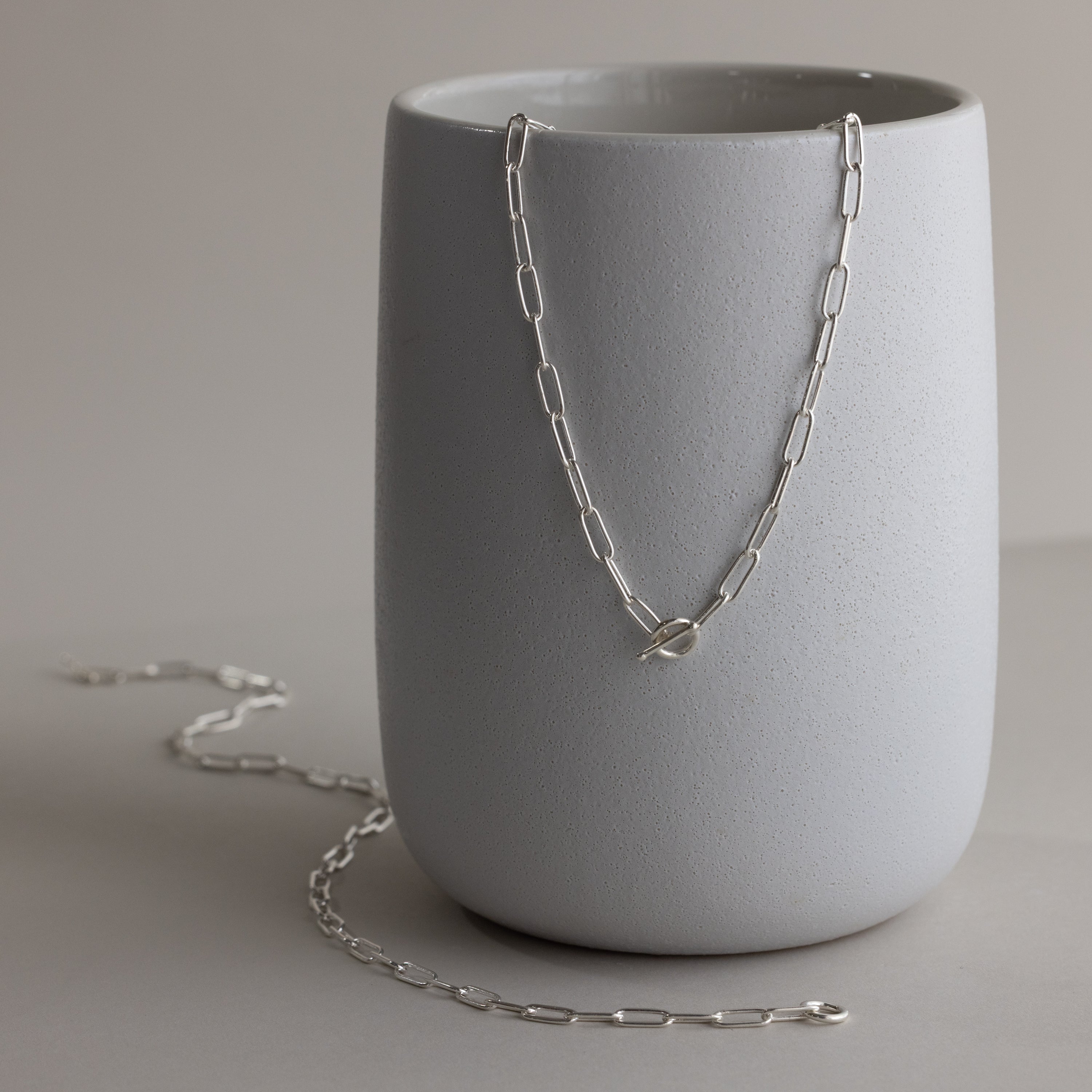 The Paperclip Toggle Necklace shines in silver atop a matte gray ceramic cup, bringing boho-chic elegance and versatility to your layered necklaces collection.