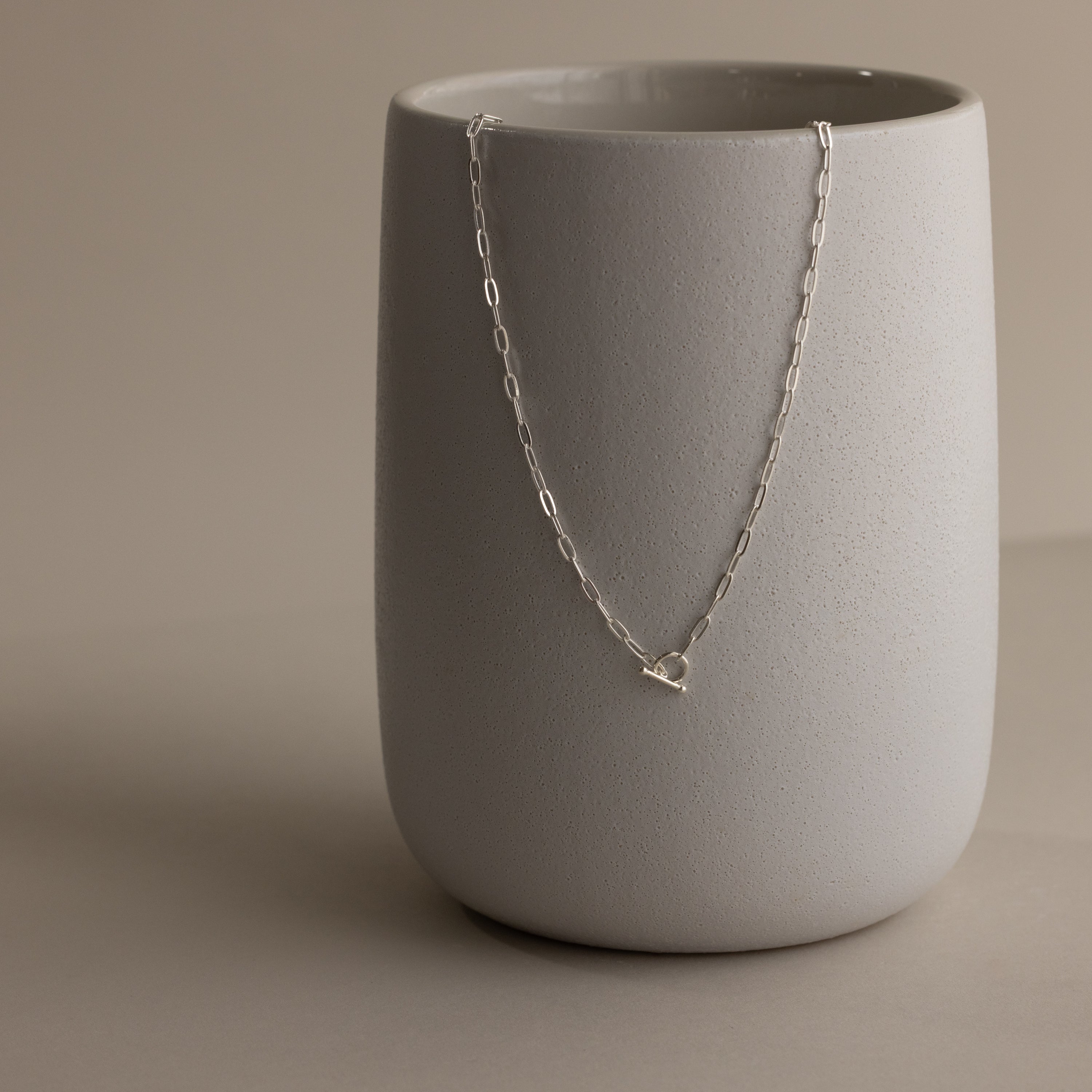 The Dainty Link Chain Necklace in Sterling Silver is draped over a matte, light gray ceramic cup on a neutral background, perfect for elegant necklace layering.