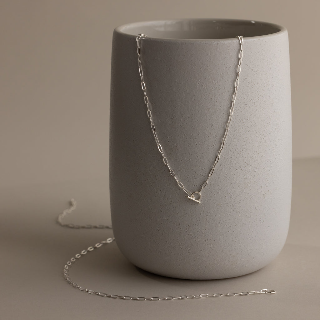The Dainty Link Chain Necklace in Sterling Silver is elegantly draped over a smooth, light gray ceramic vase on a neutral background, inspiring effortless layering.